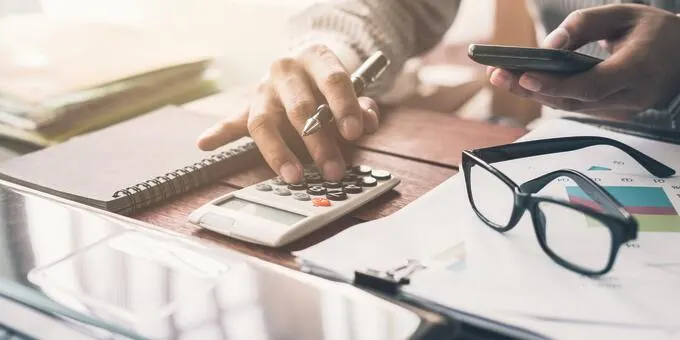 Person using calculator and phone, with glasses on a desk.