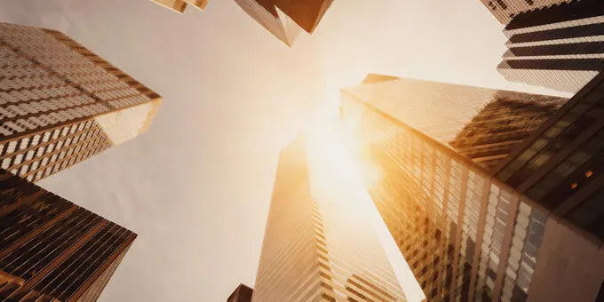 Buildings reaching into the bright sky, with sun peeking through.