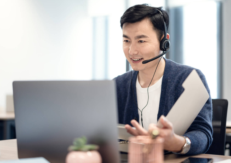 Man wearing headset, using a laptop, and holding papers.