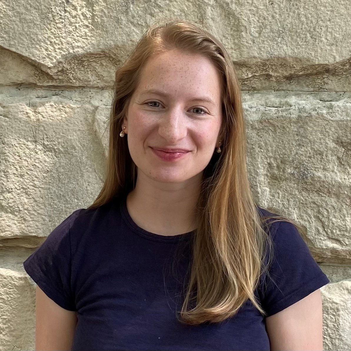 Smiling woman with long blonde hair, wearing a navy shirt, and small gold earrings, against a stone wall.