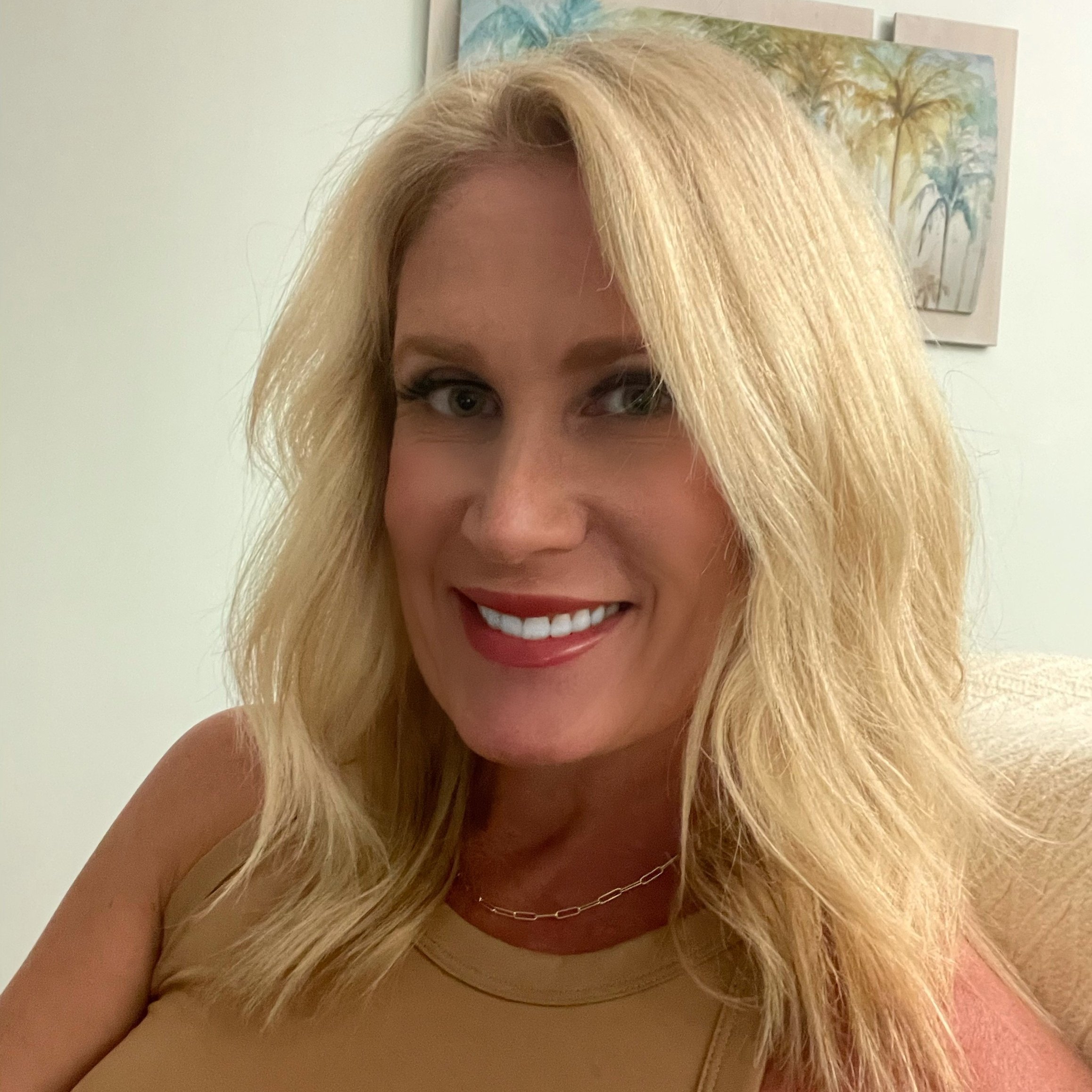 Headshot of a smiling blonde woman. She wears a gold necklace and a brown tank top. A palm tree painting hangs in the background.