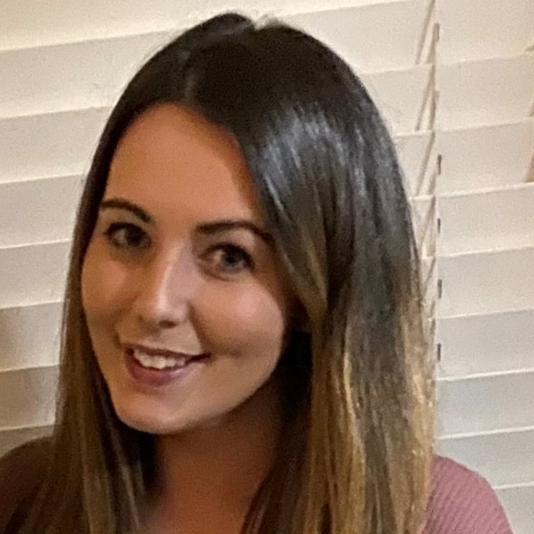 Headshot of a woman with long brown hair smiling. She is wearing a floral top and pink sweater.
