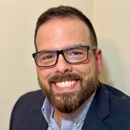 Man with beard, glasses, and blue suit jacket smiles.