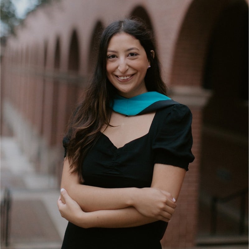 Smiling woman in black dress with arms crossed. Teal sash. Brick arches background.