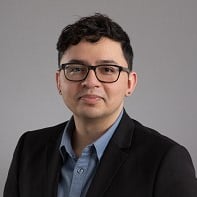 Man with glasses, black jacket, and blue shirt against gray backdrop.