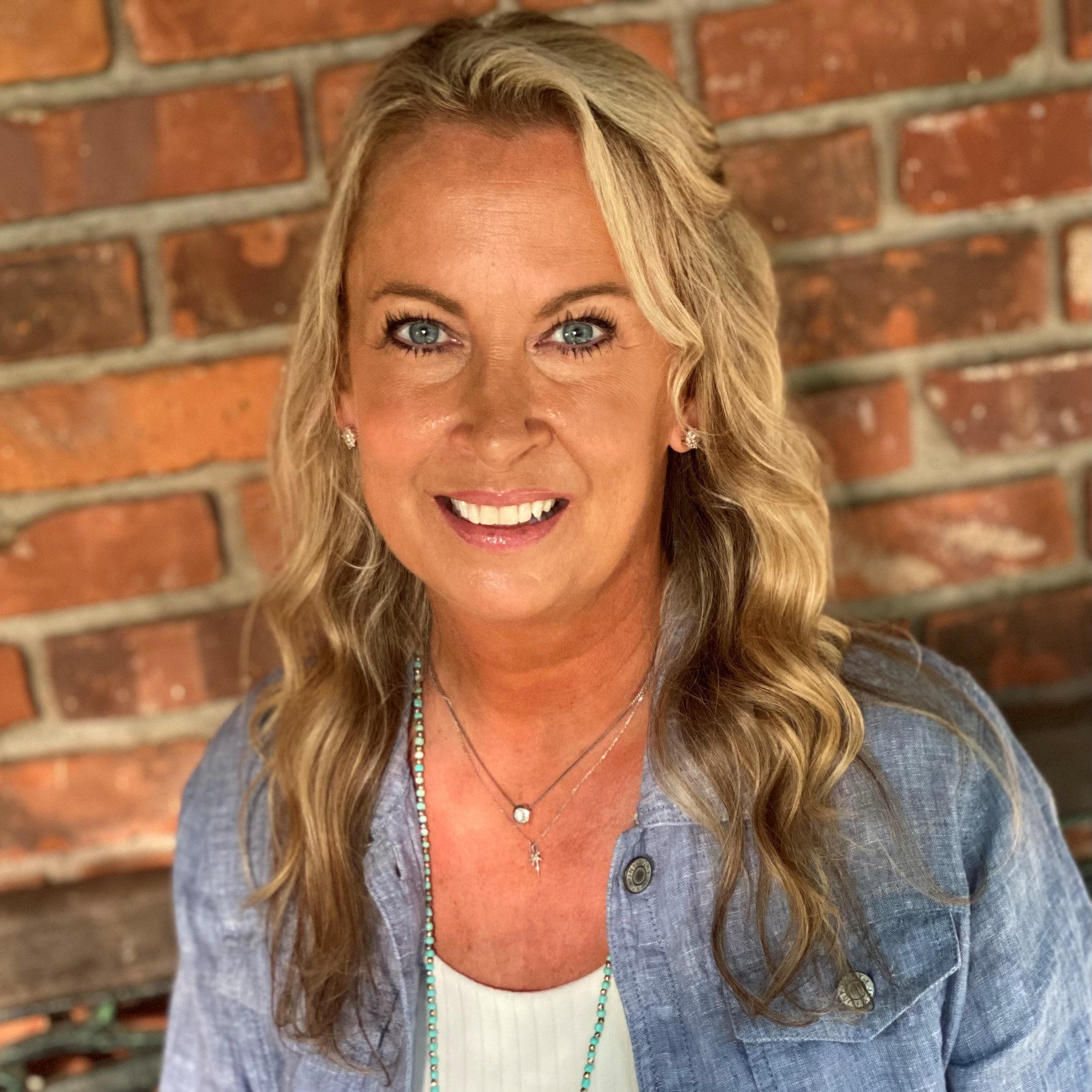 Blonde woman with blue eyes smiles at the camera, brick wall in background.
