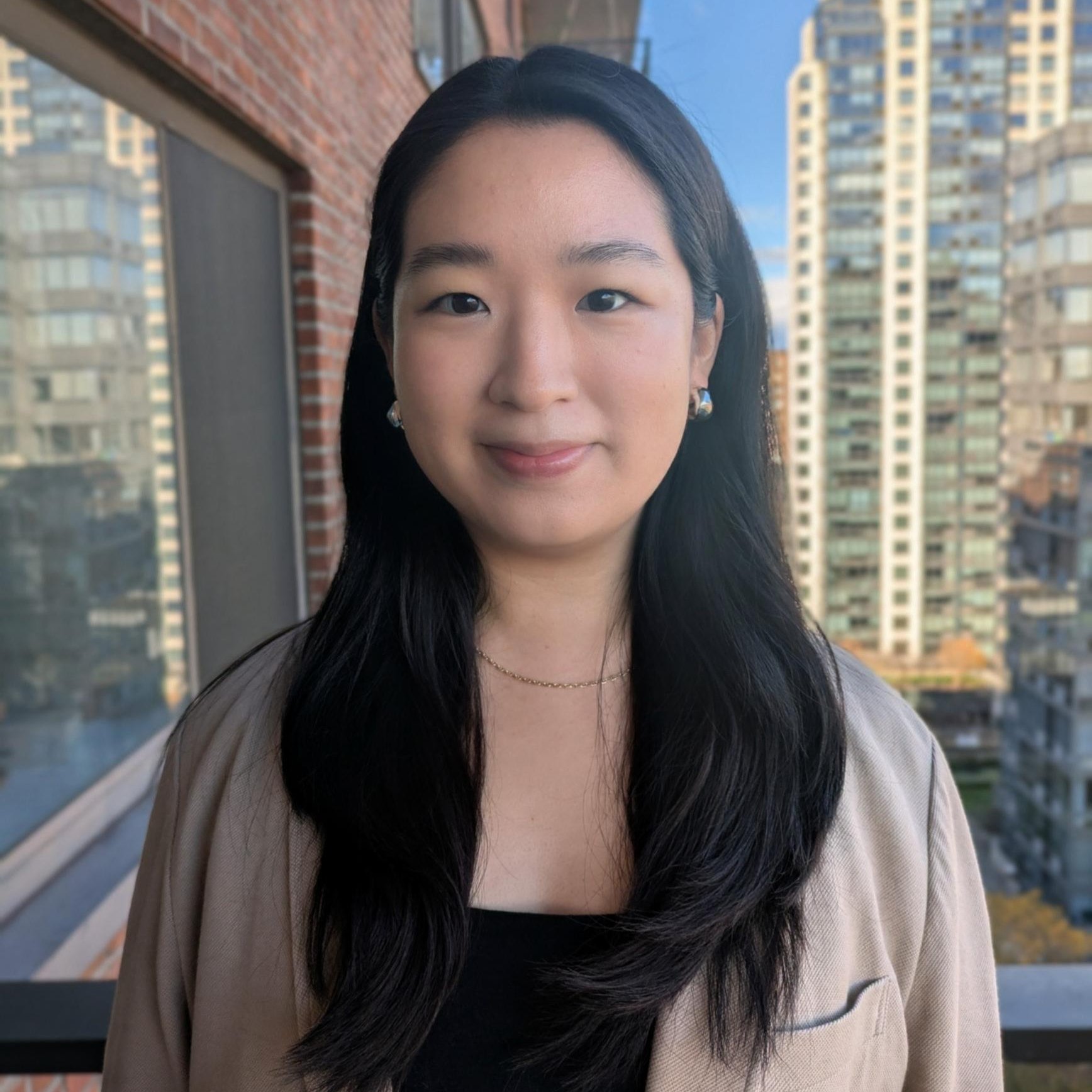 Portrait of a woman with long black hair, wearing a blazer, smiling. Buildings in the background.