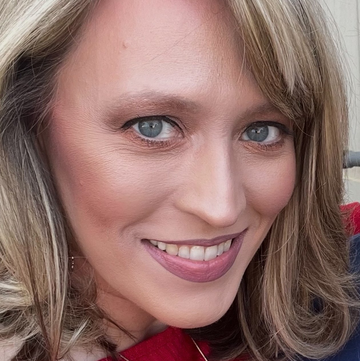 A woman with blonde and brown hair smiles directly at the camera, wearing a red top and necklace.