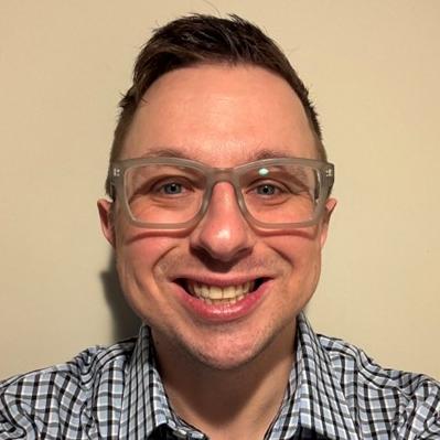 Man with blue eyes and thick-rimmed glasses smiles widely at the camera, wearing a plaid shirt.
